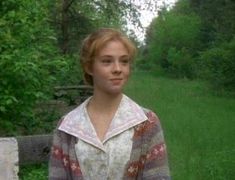 a woman standing in front of a wooden fence with trees and grass behind her, looking at the camera