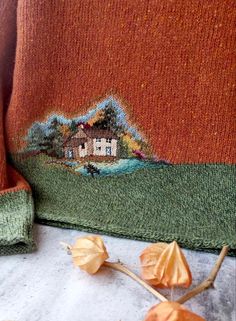 an orange sweater with a house on the front and trees in the back, next to flowers