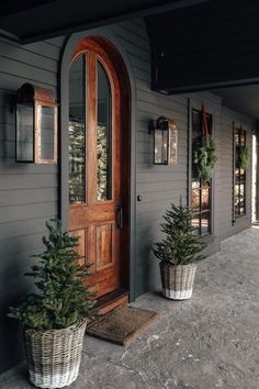 two potted trees sitting on the front porch of a gray house with wooden doors