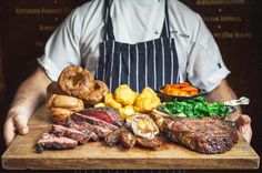 a man holding a wooden cutting board with steaks, potatoes and vegetables on it