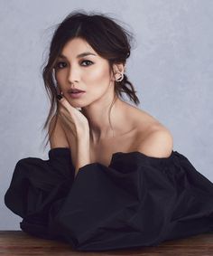 a woman sitting on top of a wooden table next to a gray wall with her hand under her chin