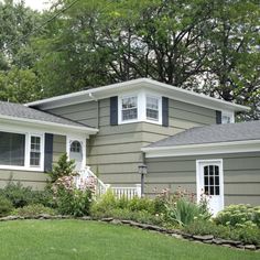 a gray house with white trim and black shutters on the windows, bushes and trees around it