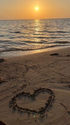 a heart drawn in the sand on a beach with the sun setting over the water