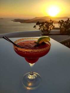 a red drink sitting on top of a table next to the ocean with a slice of lime