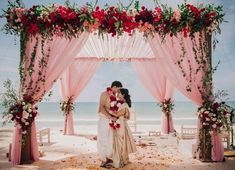 a bride and groom kissing under an archway decorated with red flowers on the beach in front of the ocean