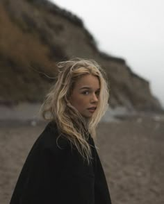 a woman standing on top of a sandy beach next to the ocean with her hair blowing in the wind