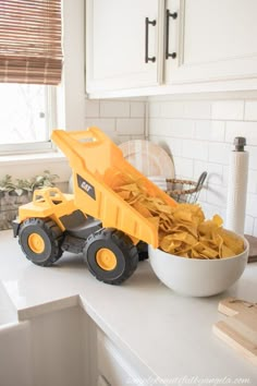 a yellow toy dump truck sitting on top of a kitchen counter next to a bowl of chips