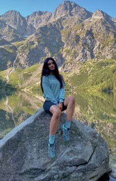 a woman sitting on top of a large rock next to a body of water with mountains in the background