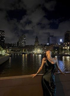 a woman in a black dress is standing on a ledge by the water at night