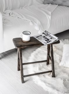 a small wooden table with a newspaper and cup on it next to a white bed