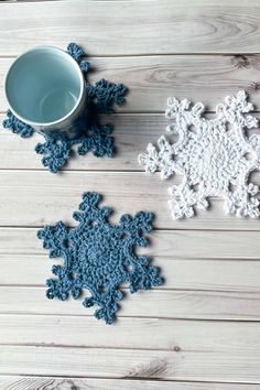 two crocheted snowflakes next to a cup on a white wooden table