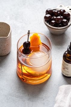 a glass filled with liquid next to two bowls full of cherries and an orange slice