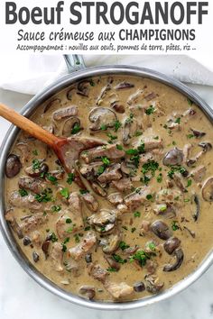 a pan filled with meat and mushrooms on top of a white table cloth next to a wooden spoon