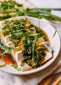 a white plate topped with tofu covered in green vegetables and broccoli next to chopsticks