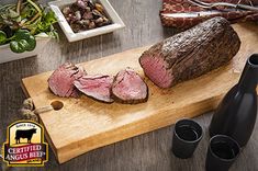 a large piece of meat sitting on top of a wooden cutting board next to two wine glasses