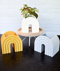 three different shaped objects on a table with a white brick wall in the background and a potted plant next to it