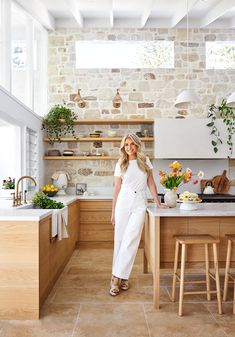 a woman standing in a kitchen next to a table with flowers on it and potted plants