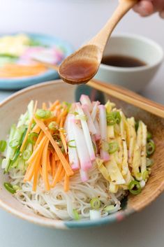 a person holding a wooden spoon over a bowl of food with noodles and vegetables on it