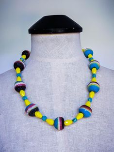 This bold oversized beaded necklace features 23mm vintage 1980s African sand cast recycled multicolor glass beads, showcasing vibrant layers of black, red, white, yellow, and blue. Complementing these striking beads are 9mm electroplated teal/blue faceted crystal coin beads, framed by 15x10mm faceted bright yellow teardrop crystal glass beads, adding an eye-catching pop of color. The collar-length design measures approximately 23 to 24.5 inches, making it a standout statement piece. The beaded s Retro Multicolor Necklaces For Jewelry Making, Retro Multicolor Round Bead Jewelry, Retro Multicolor Round Beads Jewelry, Retro Multicolor Round Beads Necklace, Retro Multicolor Round Beaded Jewelry, Retro Multicolor Jewelry For Beach, Retro Multicolor Beach Jewelry, Retro Multicolor Handmade Beaded Necklaces, Retro Multicolor Beaded Necklace