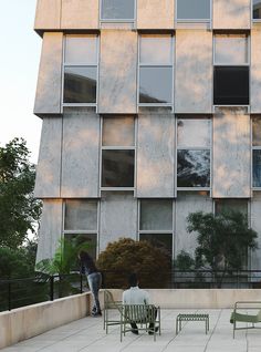 two people sitting on green chairs in front of a building with windows and balconies