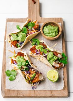 three tacos on a cutting board with guacamole and lime wedges