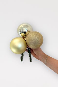 a hand is holding three ornaments in gold and silver, each with a pine branch