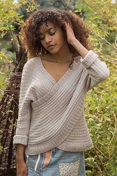 a woman with curly hair wearing a sweater and jeans in front of some bushes holding her hand on her head