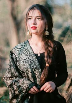 a woman with long hair wearing a black dress and gold earrings, standing in the woods