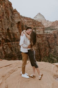 a man and woman kissing on top of a rock in the desert with mountains behind them