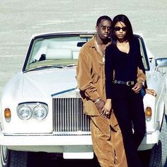 a man and woman standing next to a white car in the sand with their arms around each other