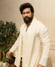 a man with a beard standing in front of a potted plant and wearing a white outfit