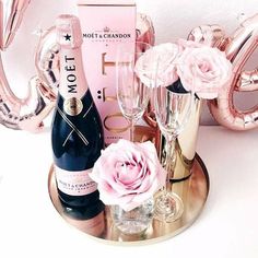 a bottle of champagne and two glasses on a tray with pink balloons in the background