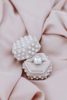 two rings with pearls on them are sitting on a pink cloth covered surface, one has a diamond in the middle and the other is white