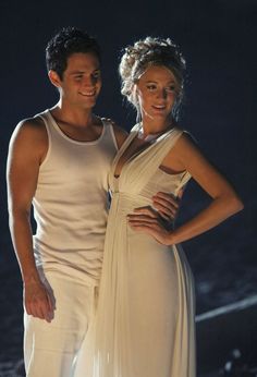 a man and woman standing next to each other in front of a dark background wearing white clothing