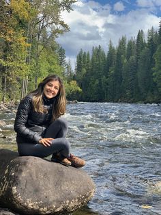 a woman sitting on top of a large rock next to a river with trees in the background