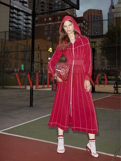 a woman standing on a tennis court wearing a red coat and white shoes with her hands in her pockets