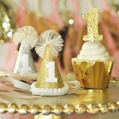 two cupcakes with gold and white decorations on top of a cake stand, one has a 1st birthday hat