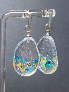 These handcrafted earrings are a true show to the artistry and creativity of their maker. Each earring features a clear resin teardrop pendant that cradles a pieces of iridescent abalone shell. The shell's natural hues ripple and shimmer within the resin, creating an enchanting play of colors that evoke the depths of the ocean. This fusion of organic beauty with the clarity of resin results in a mesmerizing and wholly unique piece of jewelry. Complementing the abalone's aquatic allure are delica Unique Teardrop Resin Earrings, Unique Iridescent Teardrop Earrings, Iridescent Abalone Shell Drop Earrings, Iridescent Dangle Resin Earrings, Blue Ocean-inspired Resin Jewelry, Abalone Shell Drop Earrings, Handmade Ocean-inspired Abalone Shell Jewelry, Bar Post, Gold Flake