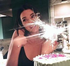 a woman holding a sparkler over a cake with white frosting and pink sprinkles