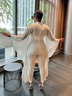 This elegant ensemble combines a sleek, form-fitting white jumpsuit with a flowing, sheer cape, creating a striking blend of modern sophistication and timeless grace. The outfit's minimalist design is accentuated by its clean lines and pure white fabric, making it perfect for high-profile events or stylish gatherings. The sheer cape adds a touch of ethereal charm, while the overall silhouette flatters the figure, offering both comfort and style. Paired with bold accessories, this outfit exudes c White Long Sleeve Jumpsuits For Evening, White Long Sleeve Jumpsuits And Rompers For Evening, Sheer Cape, Cape Jumpsuit, Outfits Minimalist, Plus Jumpsuit, Bold Accessories, Fabric Making, White Jumpsuit