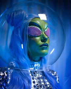 a woman with blue hair and green makeup in a bubble like space station suit, wearing pink eyeliners