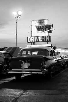 black and white photograph of old cars parked in front of a drive - in sign