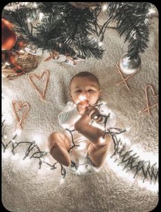 a baby sitting under a christmas tree surrounded by ornaments and lights on the floor in front of it