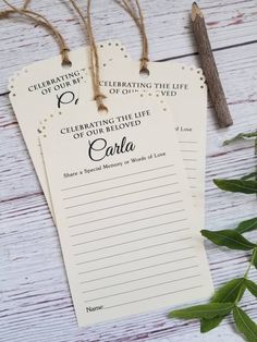 two paper tags with writing on them sitting next to some green leaves and a wooden stick