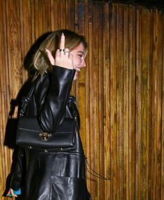 a woman wearing a black leather jacket is holding her hand up in the air while standing next to a wooden wall