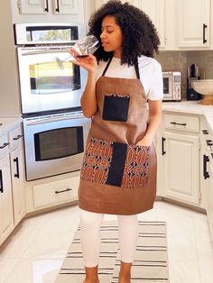 a woman standing in a kitchen with an apron on and holding a wine glass up to her mouth