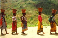 four women in colorful dresses and hats walking down a dirt road with hills in the background