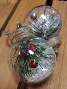 two glass ornaments with pine needles and red berries on them sitting on a wooden table