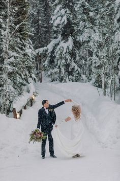 a bride and groom are standing in the snow near some trees with their arms around each other