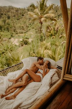 two people laying on a bed in front of a mirror and palm trees, one person kissing the other